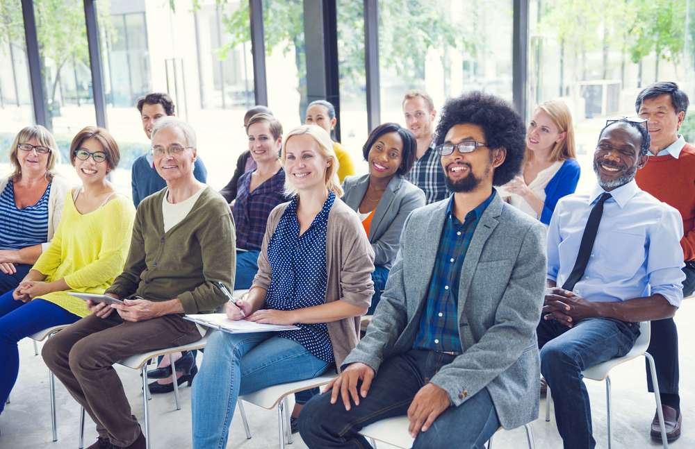 A group of adults sits on chairs in a bright room, facing forward and smiling, as though attending a seminar or presentation.