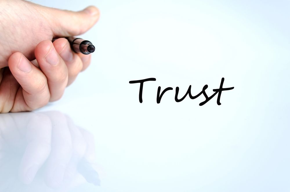 A hand holding a marker writes the word “Trust” in black ink on a white surface.