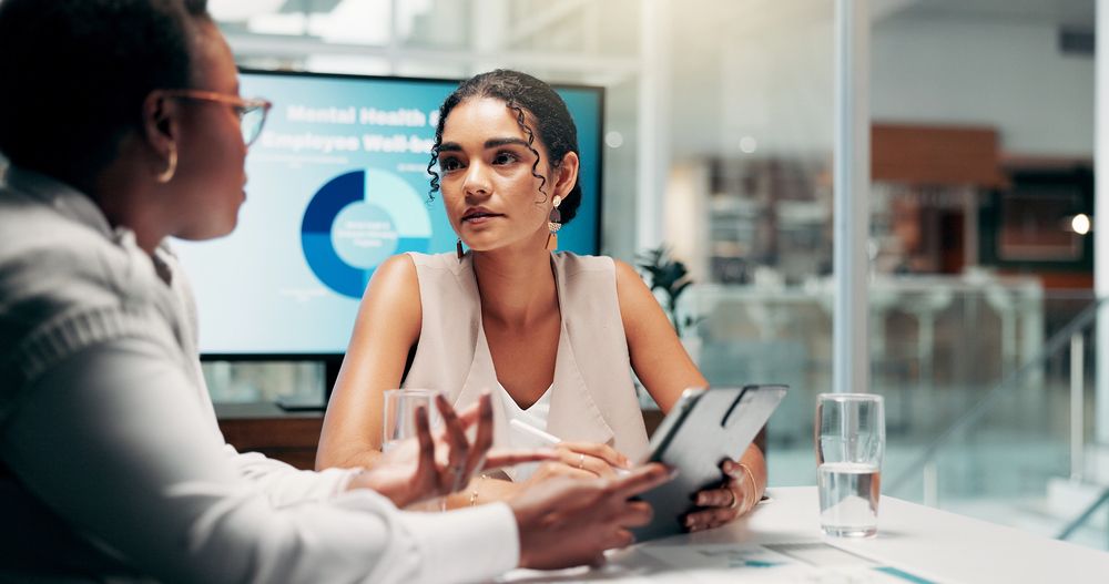 Two women sit at a desk having a discussion, with a tablet, papers, and glasses of water in front of them. An Employee Assistance Program mediator leads their conversation as a presentation on mental health is displayed in the background.